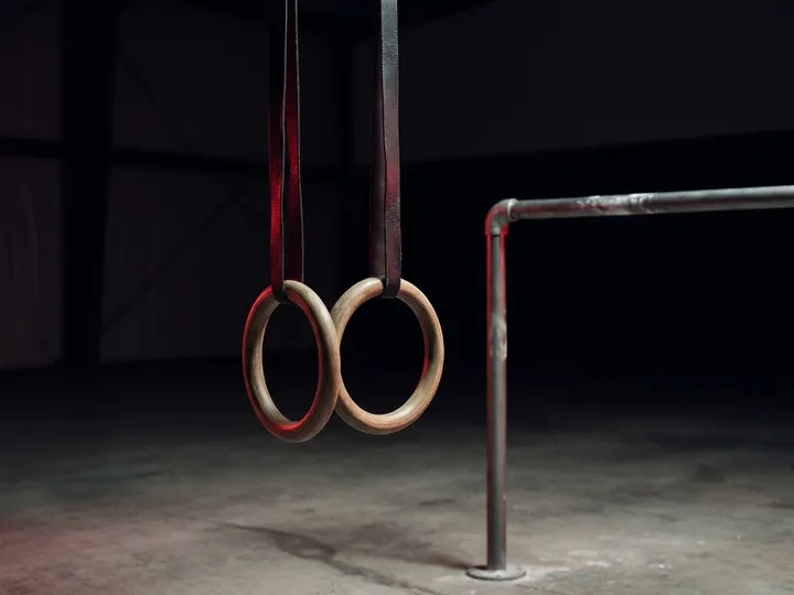Wooden gymnastic rings hanging on straps beside a steel pull-up bar in a dark training space.