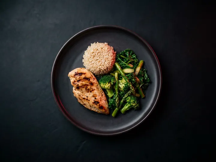 A plated meal of grilled chicken, rice and green vegetables on a dark ceramic plate, seen from above.