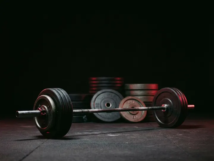 A loaded olympic barbell on the floor of a dark gym with stacked weight plates behind it.